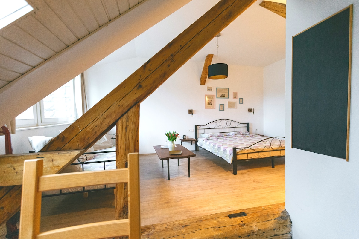 The spacious room features wooden beams and a light wooden floor. Two beds are arranged with a colorful patterned coverlet, complemented by decorative wall art. A small table is positioned in front, accompanied by a fresh bouquet of tulips. Natural light filters in through the window.