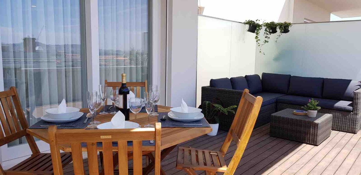 An outdoor terrace is showcased, featuring a wooden dining table set for four, complete with elegant dishware and glassware. Relaxed seating is arranged nearby with a dark, woven couch and plush cushions. The area is enhanced by greenery and offers a bright, inviting atmosphere.