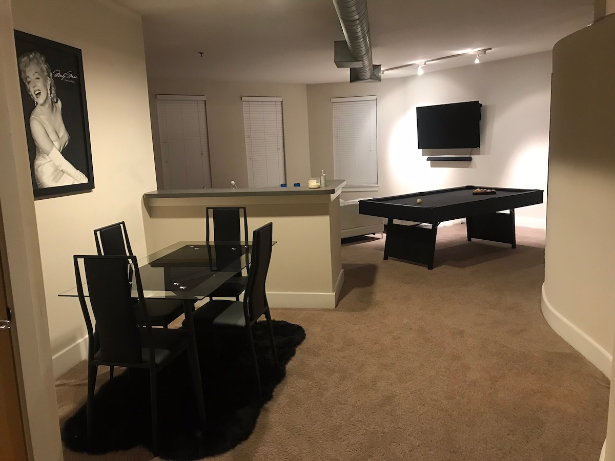 An open living area features a sleek dining table with four black chairs on a plush area rug. A modern kitchen counter is visible in the background, complemented by a pool table and a wall-mounted television, with sunlight filtering through multiple windows.