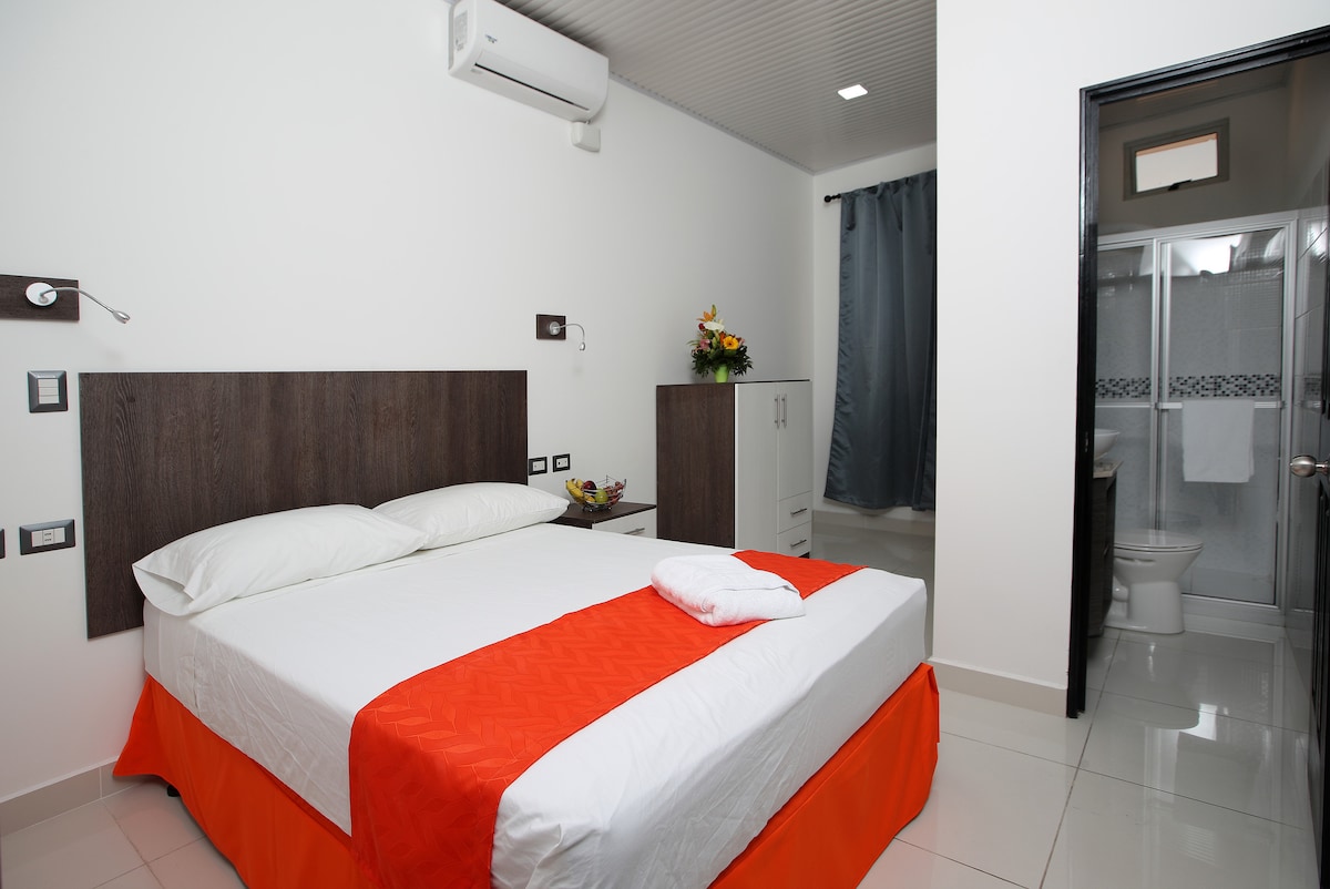A bright sleeping area features a spacious bed with a layered orange and white bedspread. The room includes a wardrobe, and a small flower arrangement decorates a nearby surface. A bathroom area is visible through an open door, offering simple design elements.