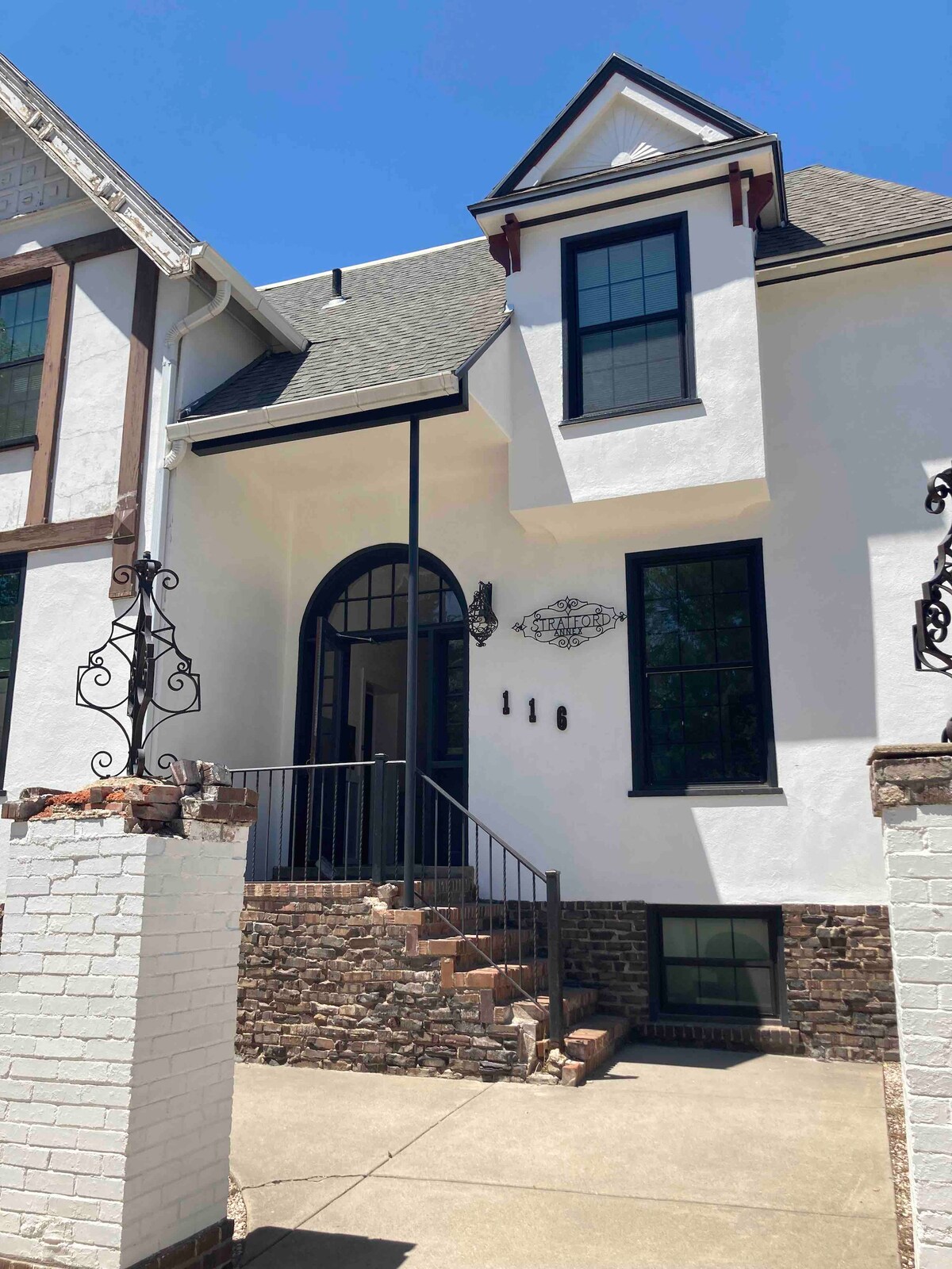 The entrance of the building showcases a white façade with stylish architectural details. A set of stairs leads to an arched doorway framed by black metal railings. The vibrant blue sky enhances the overall appearance of the entrance.