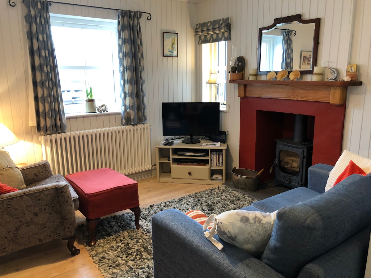 A comfortable living area features two armchairs, a coffee table, and a sofa. A wood-burning stove is set in a red-painted fireplace, with decorative items displayed above it. A mounted television is positioned on a media unit, and light filters through the window draped with patterned curtains.