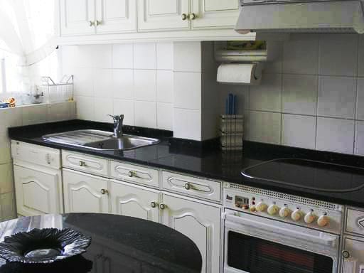 A kitchen area is presented, characterized by white cabinetry and a dark granite countertop. A stainless steel sink is situated next to an electric stove, while kitchen essentials are organized neatly. A round black plate rests atop a small island, completing the functional design.