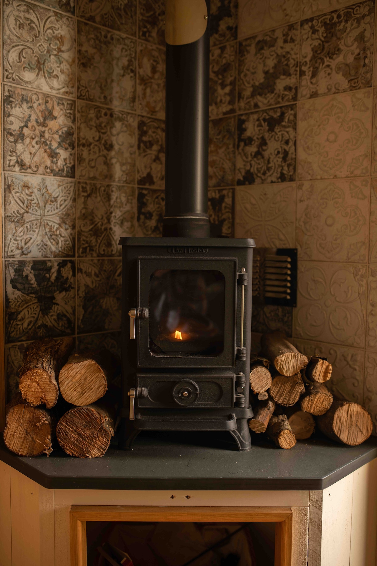 A compact wood-burning stove is positioned on a slate countertop, surrounded by neatly stacked logs. The stove features a clear glass door showcasing a glowing flame within. The backdrop consists of intricate patterned tiles in muted tones, adding texture to the space.