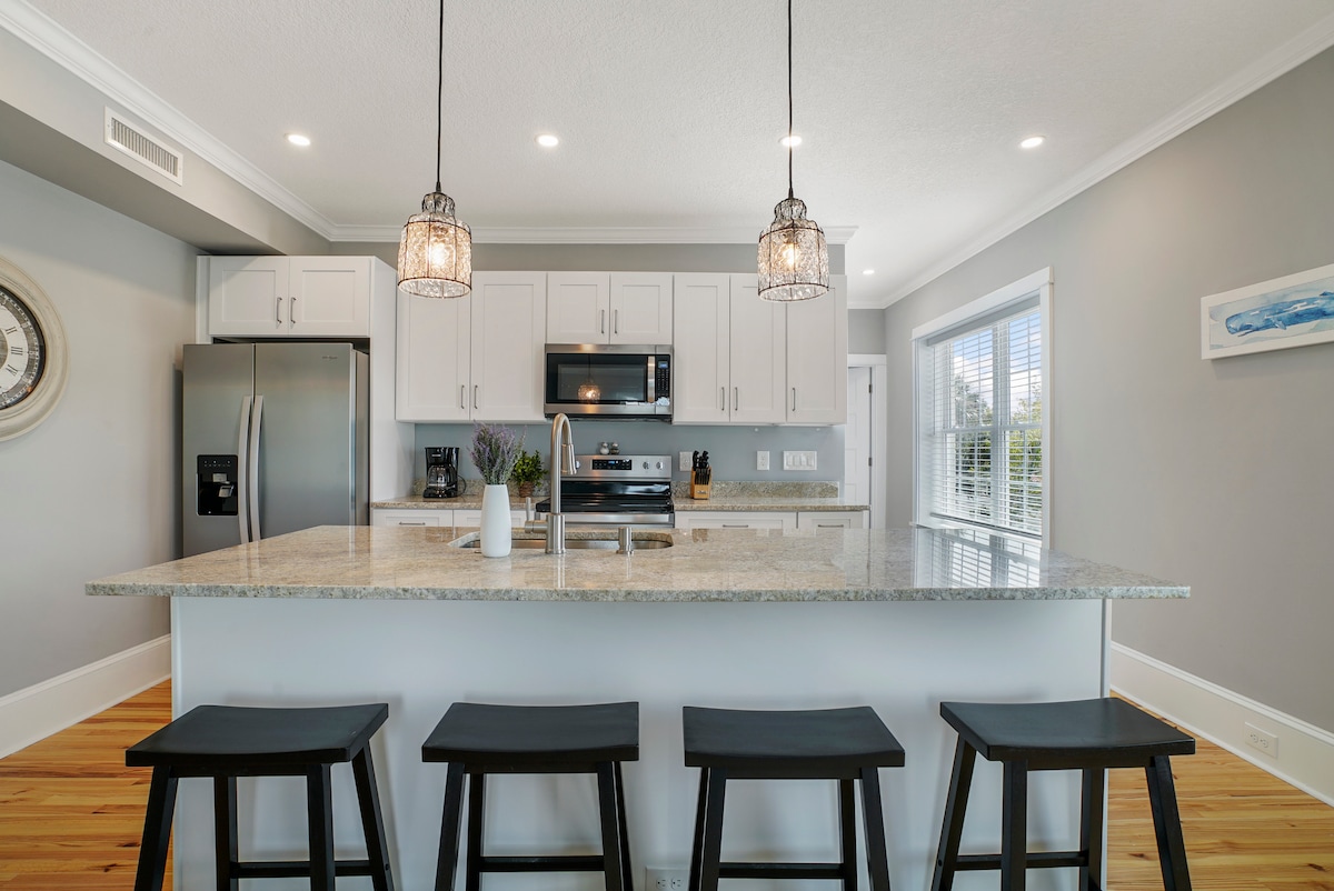 A modern kitchen showcases sleek white cabinetry, stainless steel appliances, and a large granite island with four black wooden stools. Pendant lights hang above the island, illuminating the space. Large windows provide natural light, enhancing the open and inviting atmosphere.