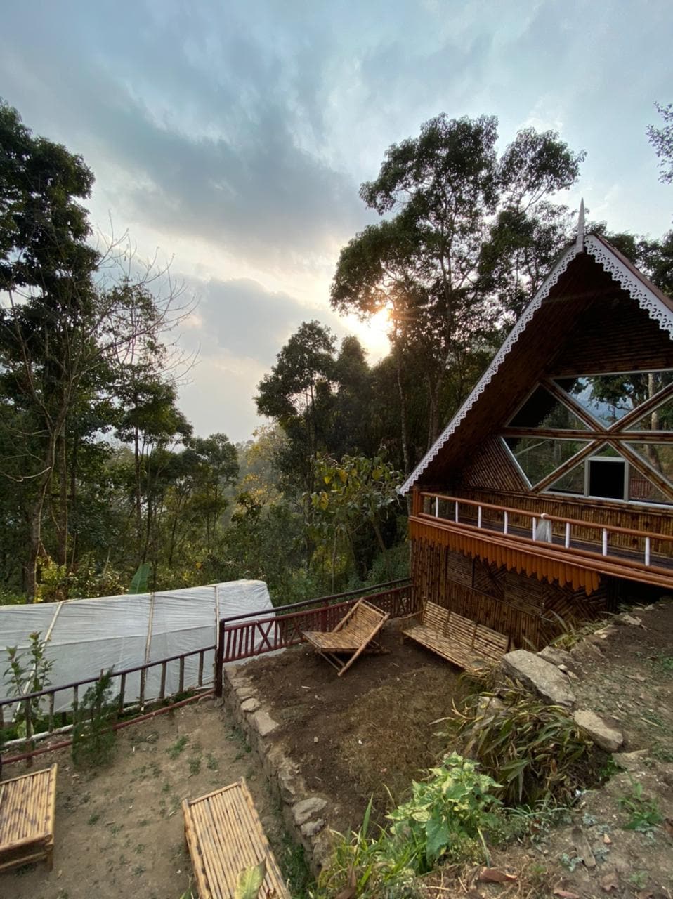 The exterior of a bamboo cottage is visible, nestled among trees. The structure features a steeply pitched roof and large windows that reflect the surrounding landscape. A small outdoor area is landscaped with bamboo furnishings arranged for seating, providing a tranquil setting with a distant view of the horizon.