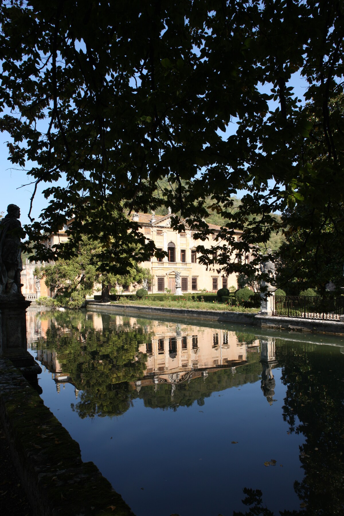 The historic Villa da Schio is reflected in the tranquil waters of a nearby canal. Surrounding trees provide shade, while marble statues are positioned along the water's edge, contributing to a serene and picturesque setting.