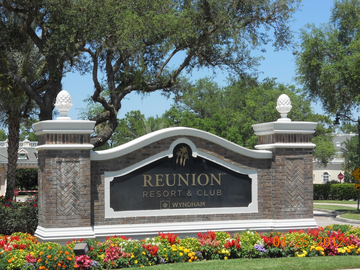 A welcoming entrance to Reunion Resort & Club is showcased, featuring a prominently displayed sign atop a decorative stone structure. Colorful flowers bloom in well-maintained landscaping, while lush greenery and trees provide a serene backdrop.