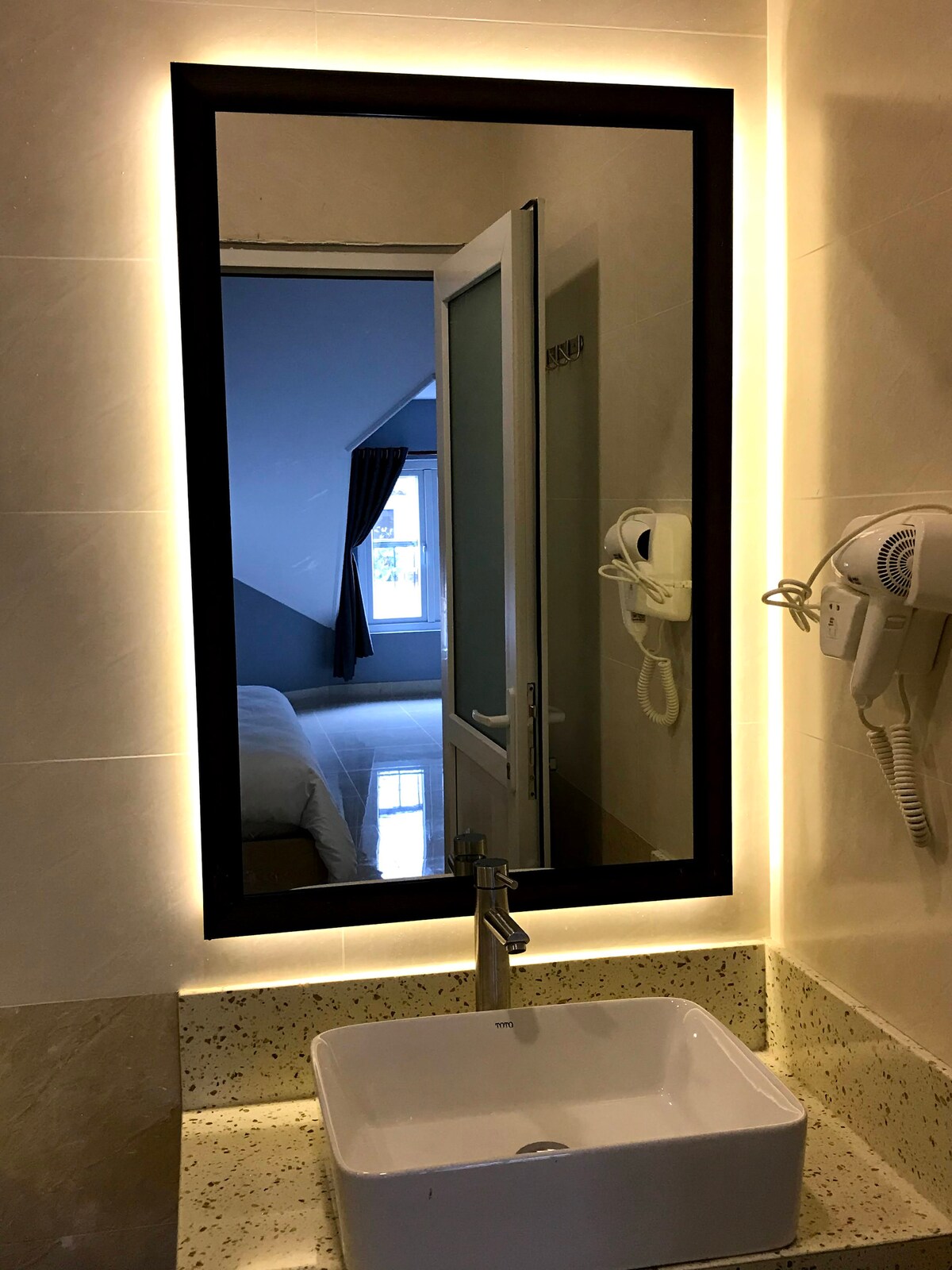 A modern bathroom sink is showcased beneath a rectangular mirror, which is framed with soft lighting. The mirror reflects a glimpse of a bedroom in the background, where natural light filters in through a window. A hairdryer is mounted beside the mirror.