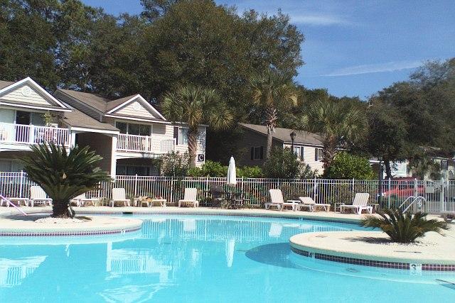 A large, inviting swimming pool features a curved design, surrounded by palm trees and lounge chairs. Nearby, two-story buildings with balconies and lush greenery create a tranquil environment. Sunlight reflects off the water, enhancing the resort-like atmosphere.