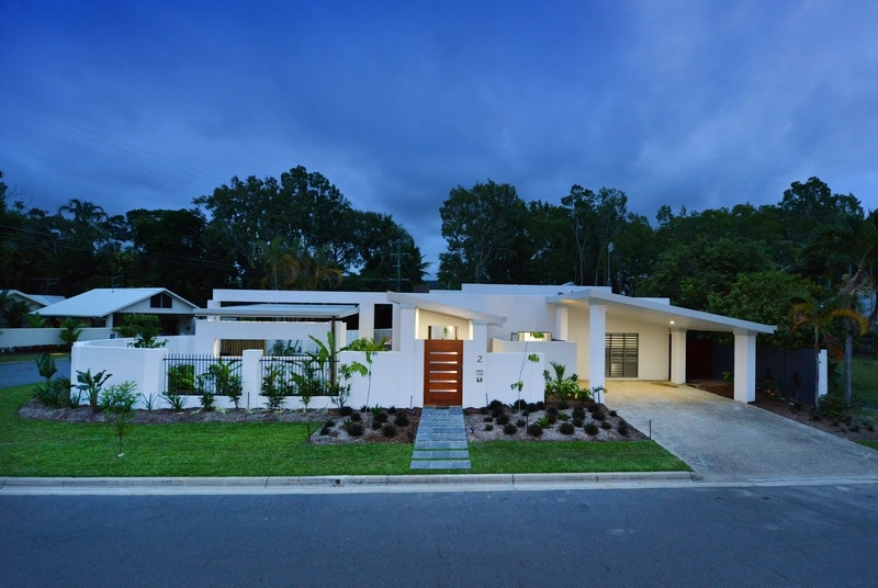 A modern beachfront property is presented with a crisp, white facade and a contrasting wooden front door. Lush greenery surrounds the entrance, while well-maintained landscaping adds to the property's inviting appearance. The driveway leads to an attached garage, framed by a serene, tropical backdrop.