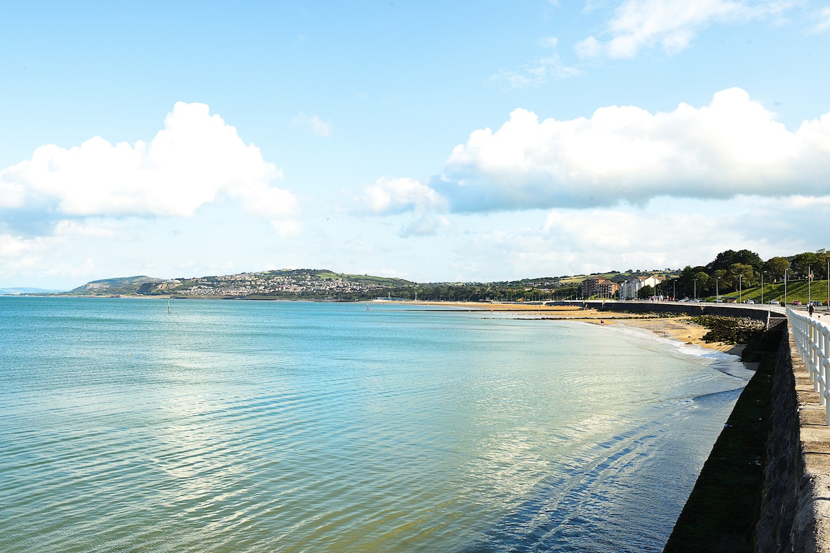 The image captures a serene beach view along the coastline. Soft waves lap against the shore, with distant hills and a coastal village visible in the background. The sky is partly cloudy, adding to the peaceful ambiance of the waterfront scene.