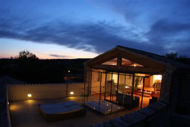 The rooftop terrace is illuminated at dusk, revealing a spacious glass structure with a wooden roof. Sun loungers are positioned for relaxation, and the surrounding views of the Luberon landscape are accentuated by the twilight sky.