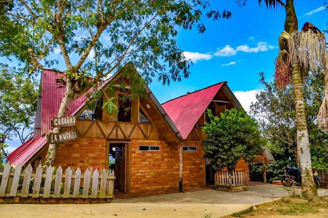 A charming cabin exterior is framed by green trees and a white picket fence. The structure features a distinctive red roof and large windows, allowing natural light to illuminate the interior. The entrance is marked by a wooden sign displaying 'Cabañas La Muralla.'