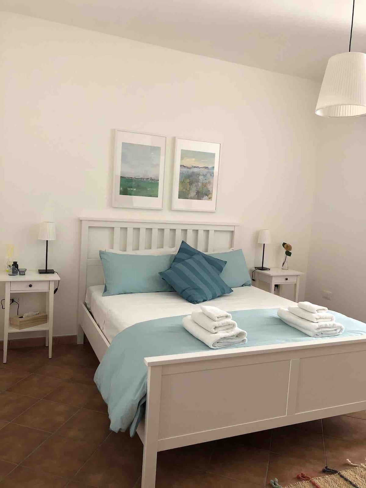 A tranquil sleeping area features a white wooden bed with blue and white decorative pillows. Two bedside tables, each with a lamp, stand on either side. Light-colored walls create a bright atmosphere, while simple artwork hangs above the bed.