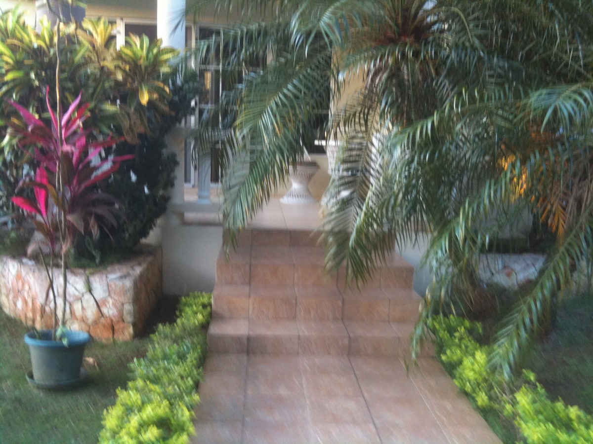 A series of stone steps leads to an entrance, flanked by lush greenery and colorful tropical plants. The pathway is bordered by well-maintained grass and emerges from a vibrant garden setting.