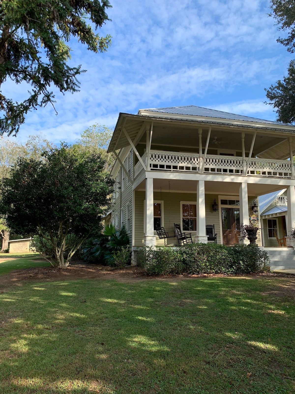 A charming historic carriage house is set amidst lush greenery. The porch features rocking chairs, while a shaded yard offers ample space for relaxation. A large tree provides additional shade, enhancing the serene outdoor environment. The sky is clear with a few wispy clouds.