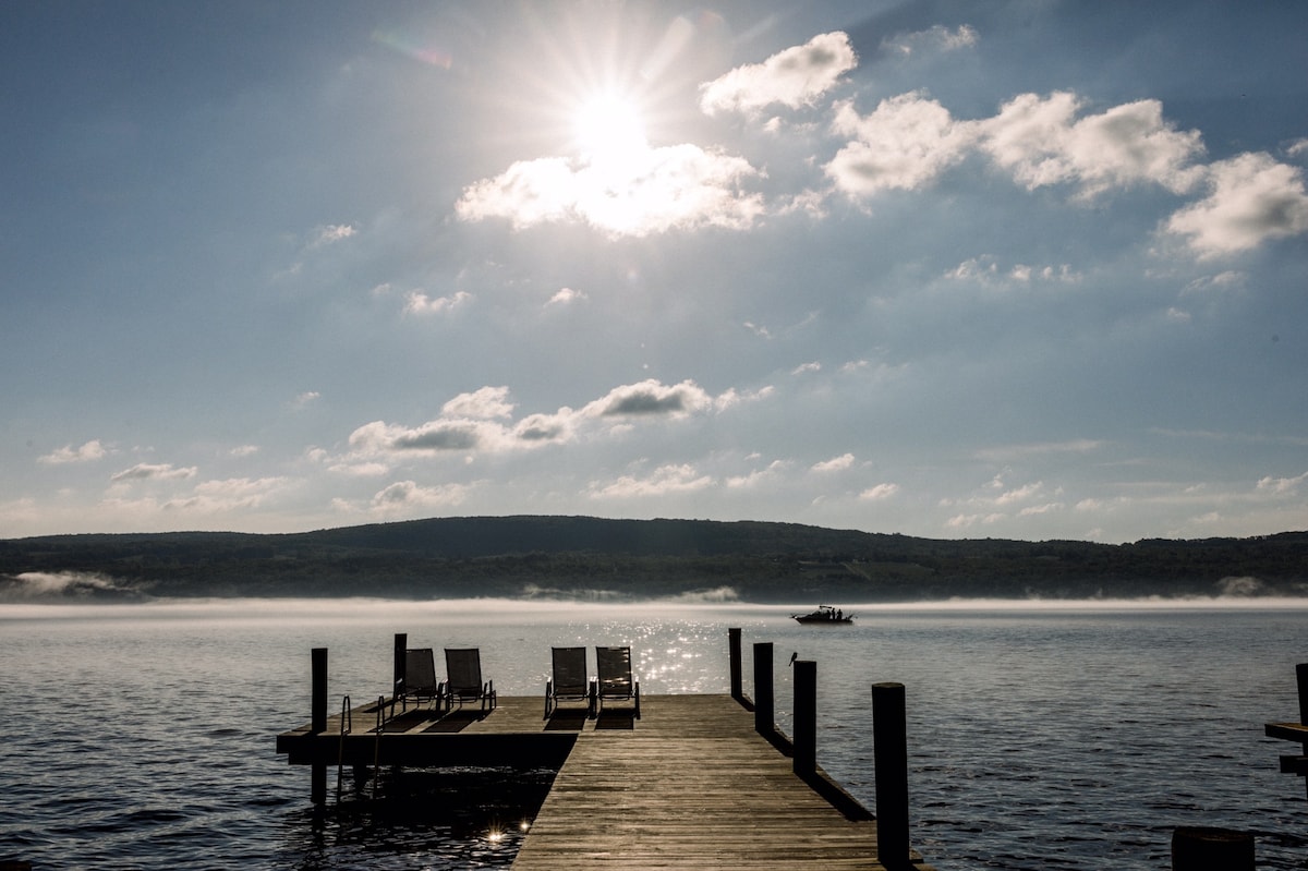 A wooden dock extends over the lake, featuring several sun loungers positioned for relaxation. The sun is seen shining brightly overhead, casting light across the tranquil water and distant shoreline, contributing to a serene outdoor setting.