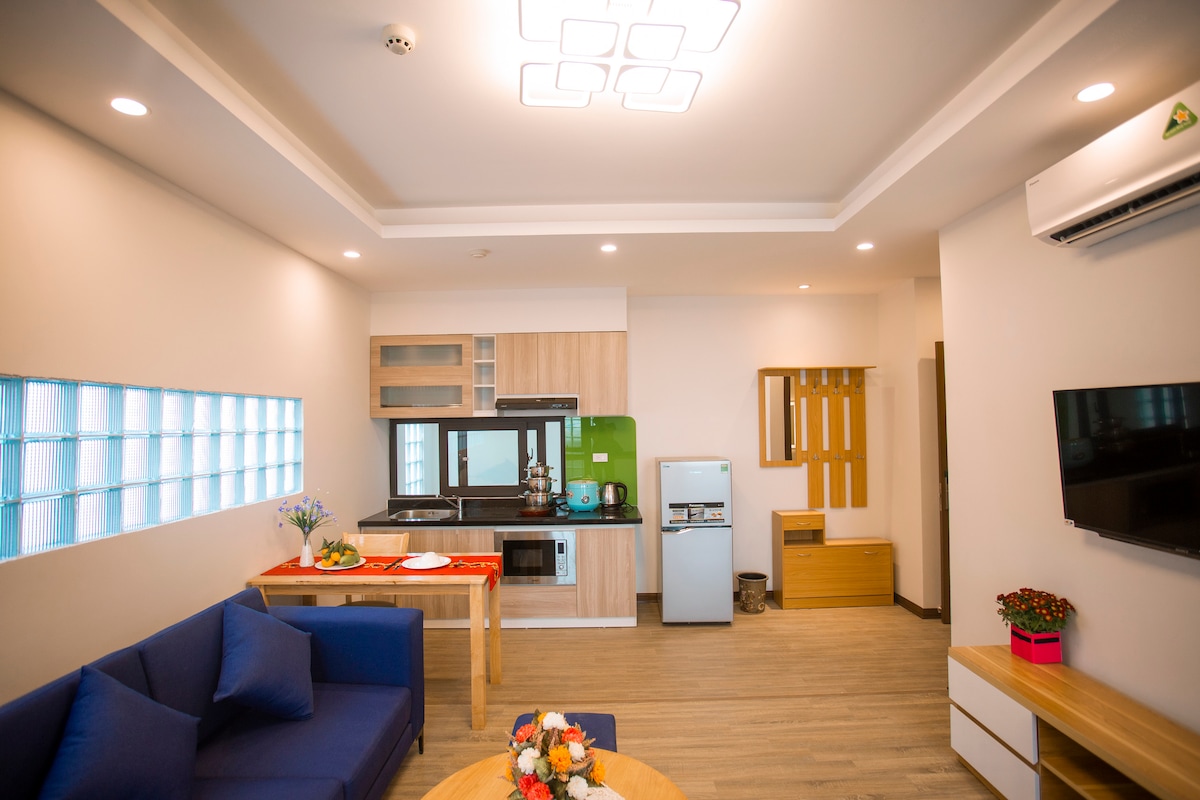 A modern living space features a blue sofa and a dining table. The kitchenette is equipped with appliances and a green accent wall. A small window allows light to enter, while decorative plants add a touch of color. A television is mounted on the wall.