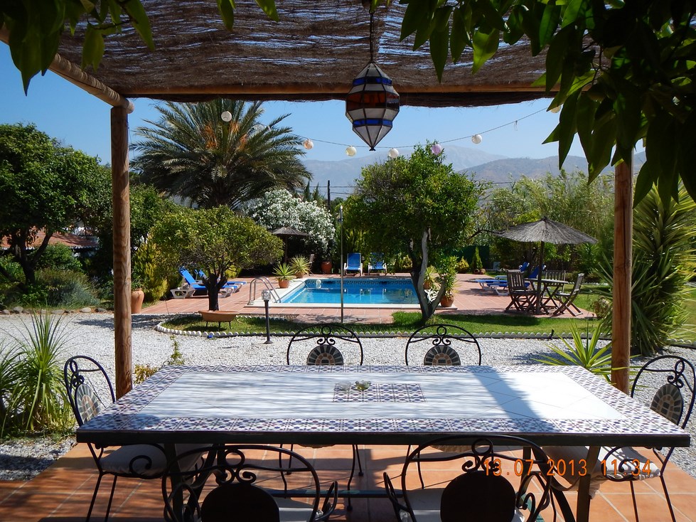 A spacious outdoor dining area is furnished with a large table surrounded by chairs, offering a view of lush gardens and a swimming pool. Sun loungers and a shaded umbrella are visible nearby, framed by mature trees and vibrant greenery.