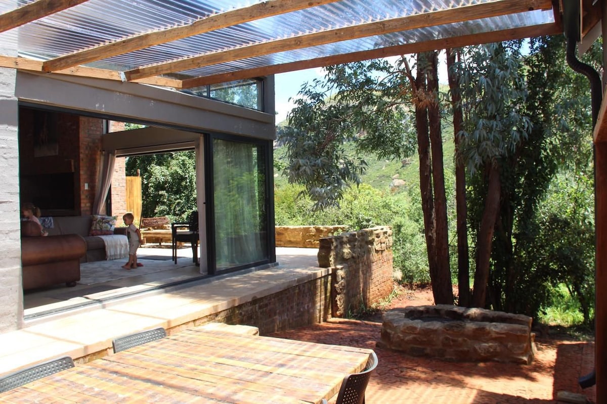 An outdoor seating area is presented under a covered patio, featuring a large table and comfortable chairs. Beyond, lush greenery and a stone fire pit create a peaceful setting. Large glass doors open to an inviting interior space, allowing natural light to fill the area.