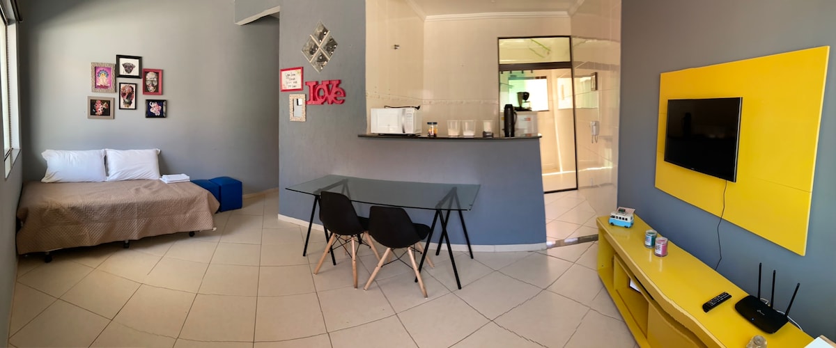 An open living area features a spacious sofa bed and a dining table with black chairs. A wall-mounted TV is positioned above a colorful yellow TV unit. The kitchenette is visible in the background, with modern appliances and a neutral color scheme enhancing the space.