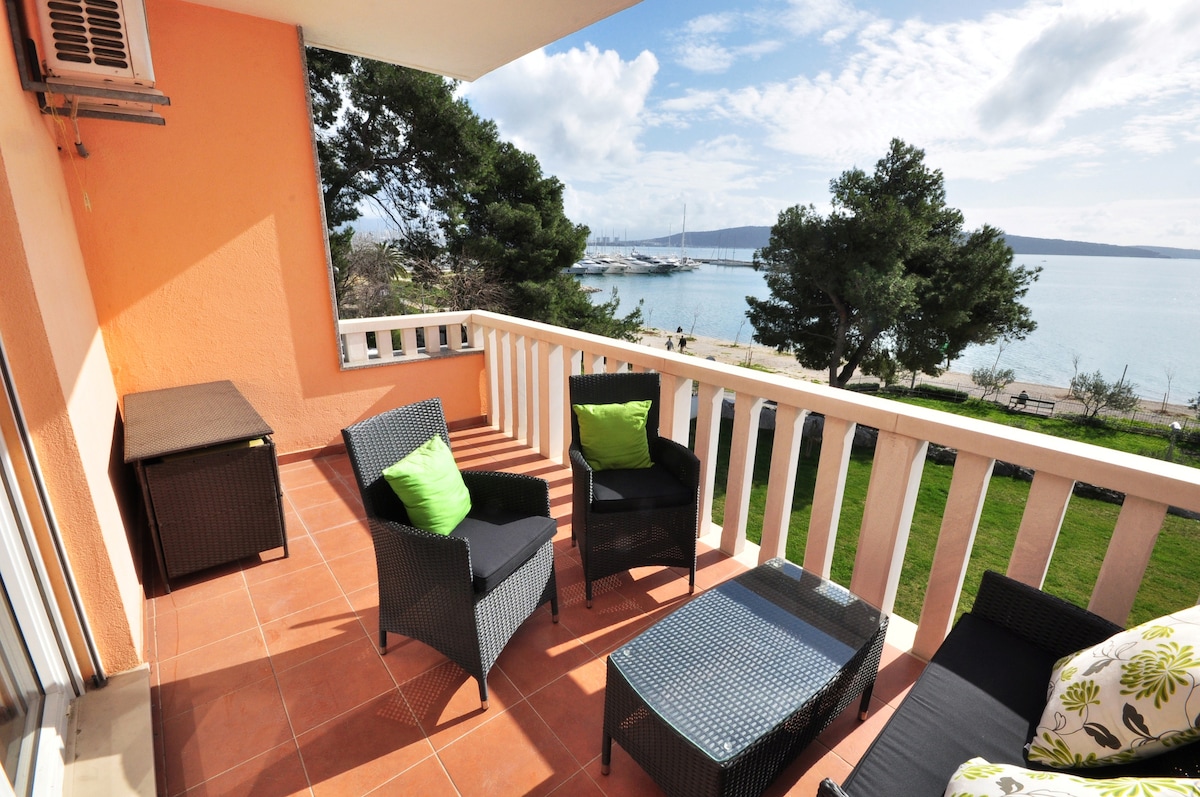A furnished balcony overlooks a serene beach and calm sea, with comfortable seating featuring green cushions. Slanted umbrellas provide shade, while a small table complements the seating area. Large windows allow for natural light, enhancing the outdoor experience.