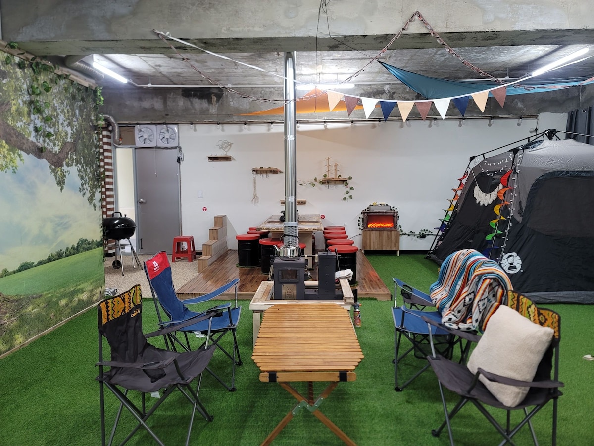 The indoor camping-inspired space features a cozy seating area with multiple canvas chairs arranged around a wooden table. A portable fireplace is positioned centrally, complemented by a decorative tent and a backdrop of nature-themed art. Colorful bunting adds a festive touch overhead.