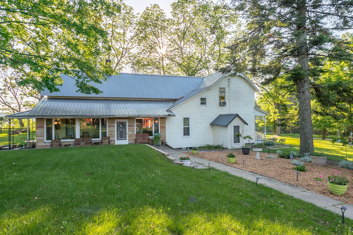 A spacious Victorian farmhouse is surrounded by lush greenery, featuring a metal roof and multiple entrances. Large windows allow natural light to filter into the interior areas. Flower pots and garden pathways guide visitors through the well-maintained outdoor space.