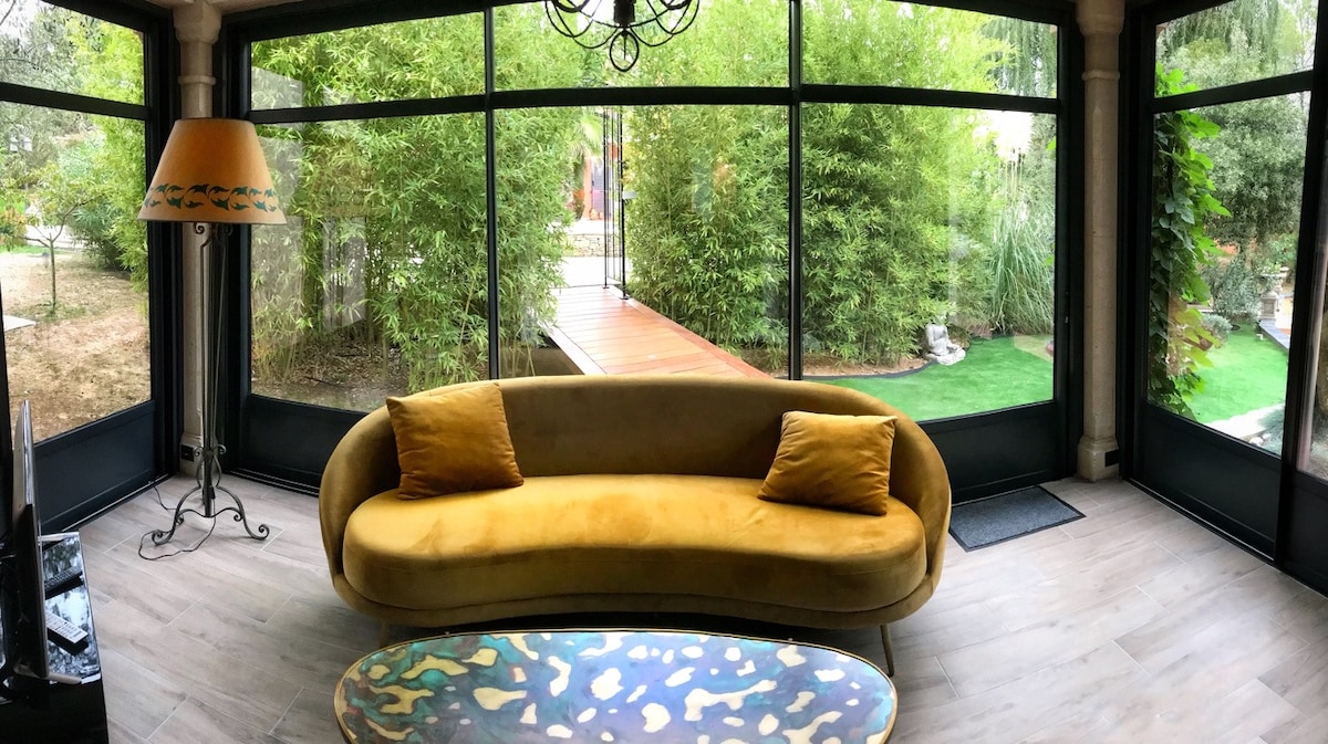 A cozy seating area features a curved yellow sofa, complemented by two decorative cushions. A colorful coffee table is placed in front, and large glass windows provide a view of the lush green garden outside, creating a bright and airy atmosphere.