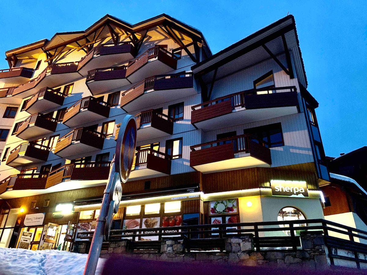 The exterior of a multi-story building is captured at dusk, featuring wooden balconies and large windows. The ground level houses a store named Sherpa, while snow is visible on the ground, reflecting the winter setting. A street sign is positioned in front of the structure.