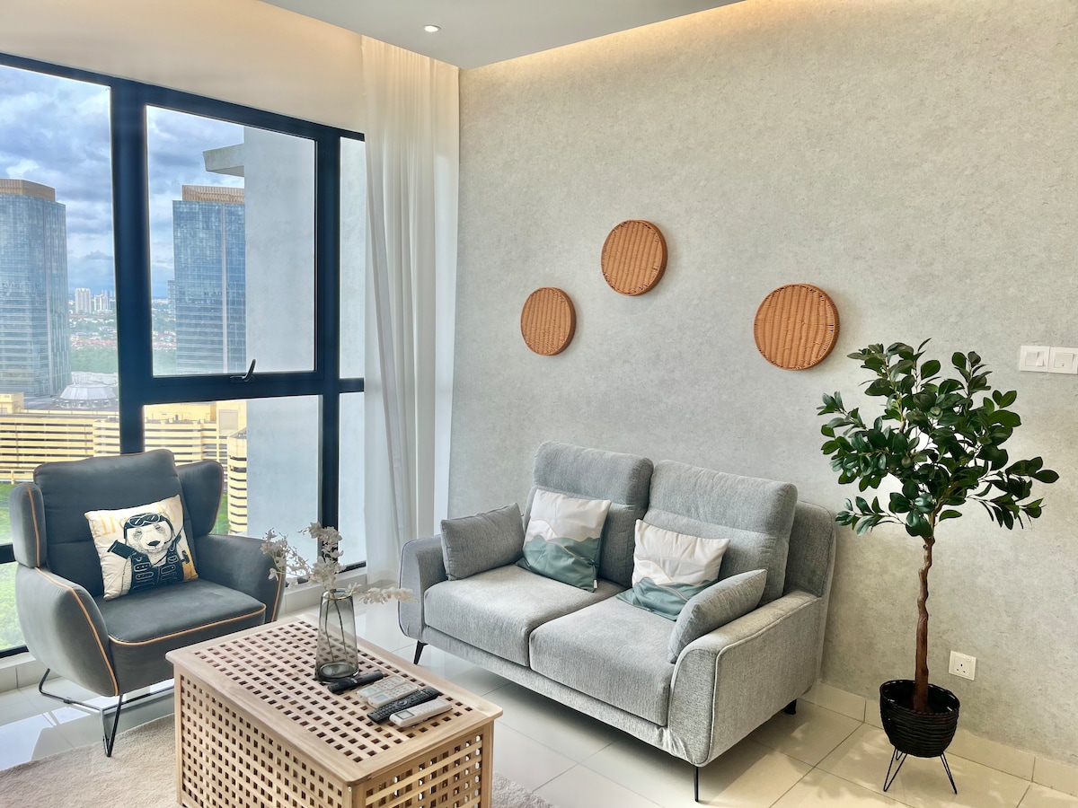 The living area features a light grey sofa paired with an accent chair, both positioned near a large window offering a city view. Three circular wooden wall decorations are displayed above the sofa. A small coffee table rests in the center, surrounded by decorative elements.