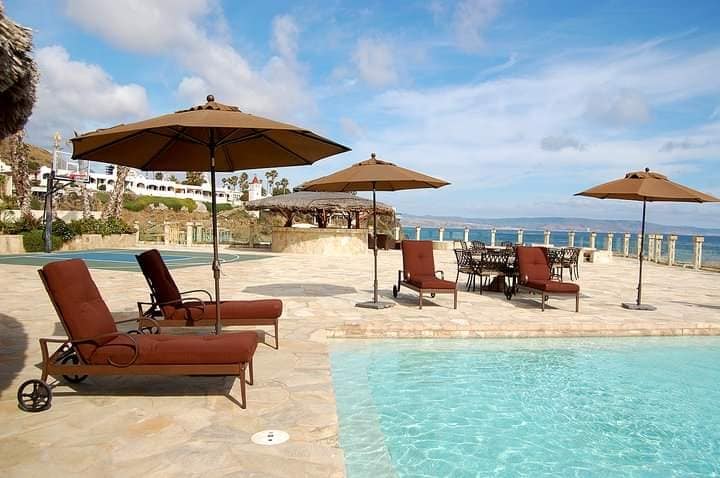 An outdoor area features a clear swimming pool surrounded by sun loungers and umbrella shades. Additional tables and chairs are arranged nearby, with scenic views of the ocean and distant hills visible under a partly cloudy sky.