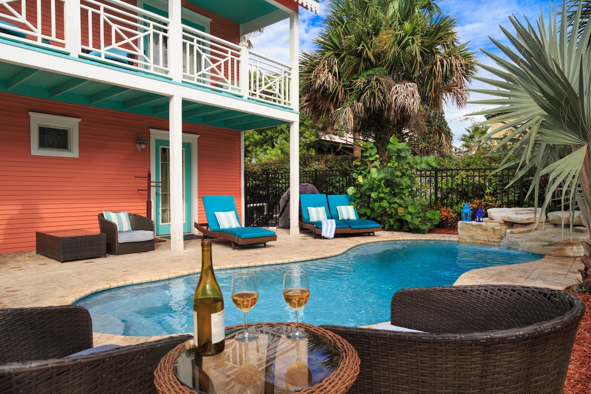 A private pool with a waterfall is surrounded by tropical landscaping and stone accents. Lounge chairs in turquoise invite relaxation, while a small table set with two glasses and a bottle of wine offers a cozy seating area. The house features an inviting coral exterior.