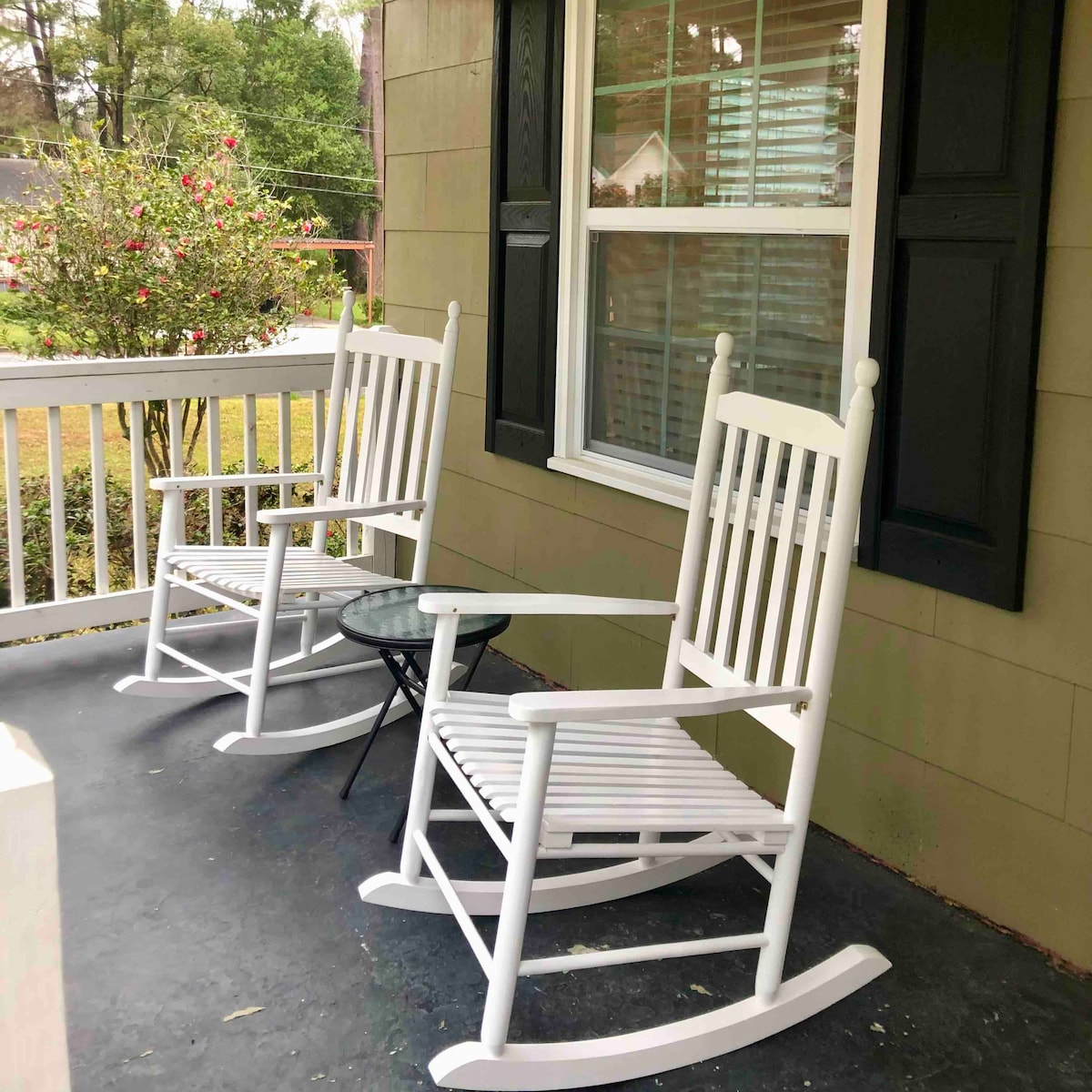 Two white rocking chairs are positioned on a front porch, facing a small round table. A bush with blooming flowers is visible in the background, and a window with shutters is seen alongside the seating area, creating a welcoming outdoor space.