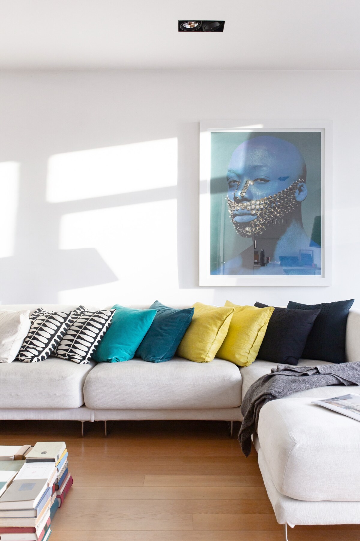 A modern living area features a light-colored sectional sofa adorned with colorful cushions in shades of teal, yellow, and black. A framed artwork is displayed on the wall. Natural light illuminates the space, highlighting the warm wooden flooring beneath.