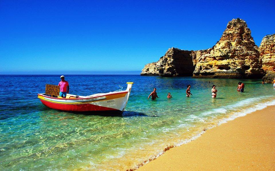 A vibrant beach scene features clear blue waters lapping against golden sands. A wooden boat with a colorful trim is anchored nearby, while individuals enjoy the refreshing water. Rock formations rise majestically in the background under a bright, clear sky.