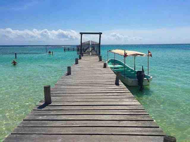 A wooden pier extends over clear turquoise water, leading to a small boat with a canopy. Calm waves reflect the sunny sky, while a few figures can be seen swimming in the distance. The horizon is dotted with soft clouds, enhancing the serene coastal setting.