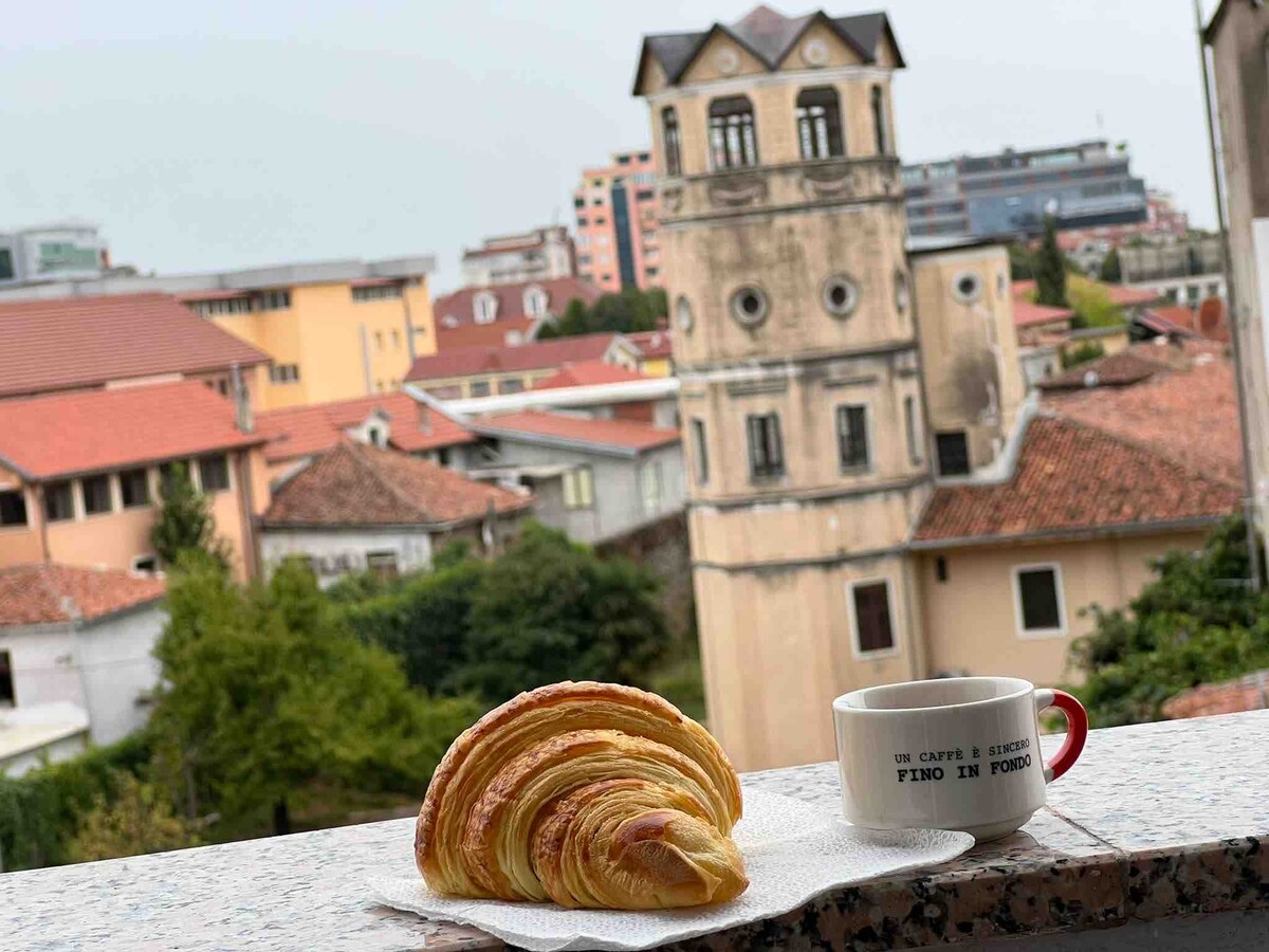 A balcony view displays a croissant and a coffee cup, with a historical tower and city rooftops visible in the background. Soft, muted colors dominate the scene, creating a peaceful atmosphere. The arrangement invites guests to enjoy their breakfast while taking in the surrounding urban landscape.