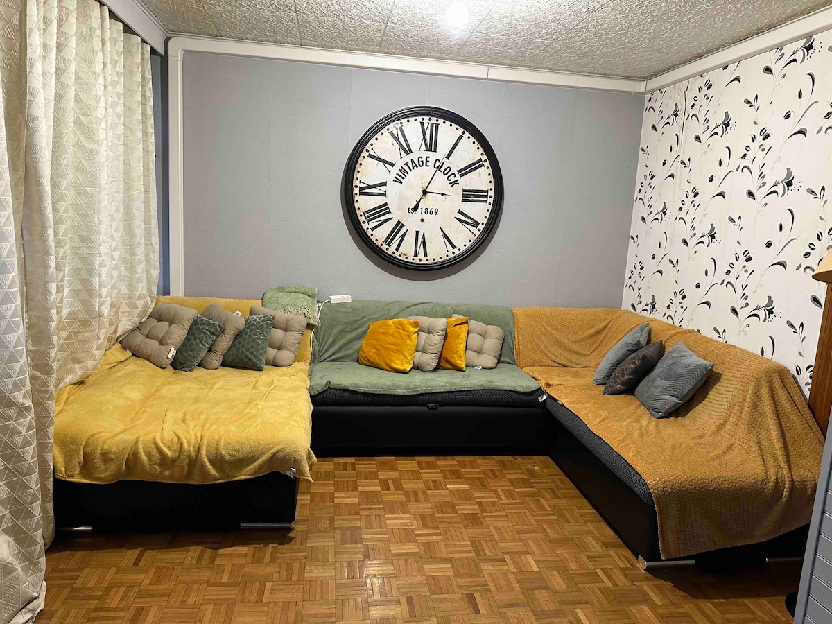A spacious living area is furnished with a large sectional sofa covered in various blankets and cushions. A decorative wall clock is prominently displayed above the sofa. The room features light-colored curtains and a patterned wallpaper, enhancing the overall ambiance.