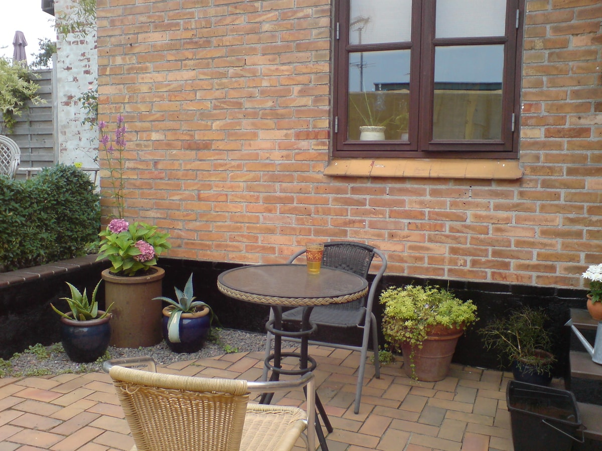 A small outdoor seating area is displayed, featuring a round table with two chairs. Potted plants, including colorful flowers and green foliage, are positioned around the space. A brick wall in the background adds texture, and a window above enhances natural light.