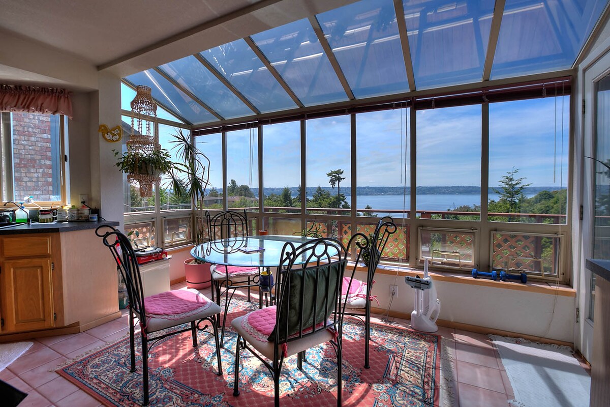 A sunlit dining area features a glass ceiling, allowing natural light to fill the space. A circular table is surrounded by six chairs, creating a welcoming atmosphere. The expansive windows showcase views of Commencement Bay, enhancing the serene environment.