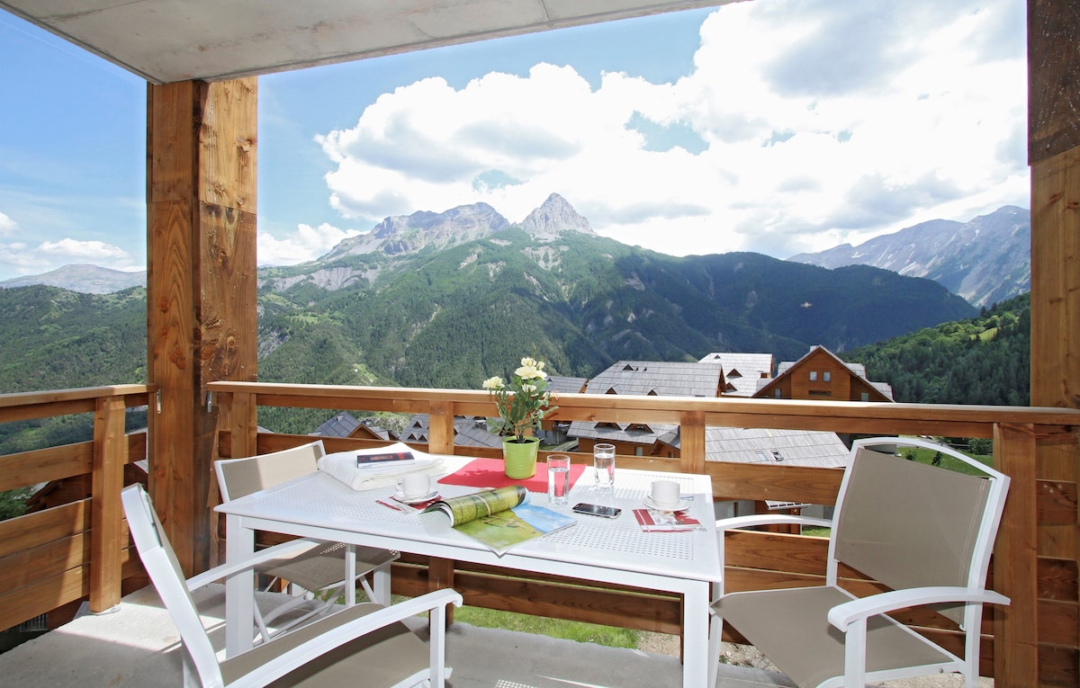 A balcony is presented with a white outdoor dining table set for four, featuring a plant centerpiece. The view showcases rolling green mountains under a partly cloudy sky, creating a peaceful natural backdrop.