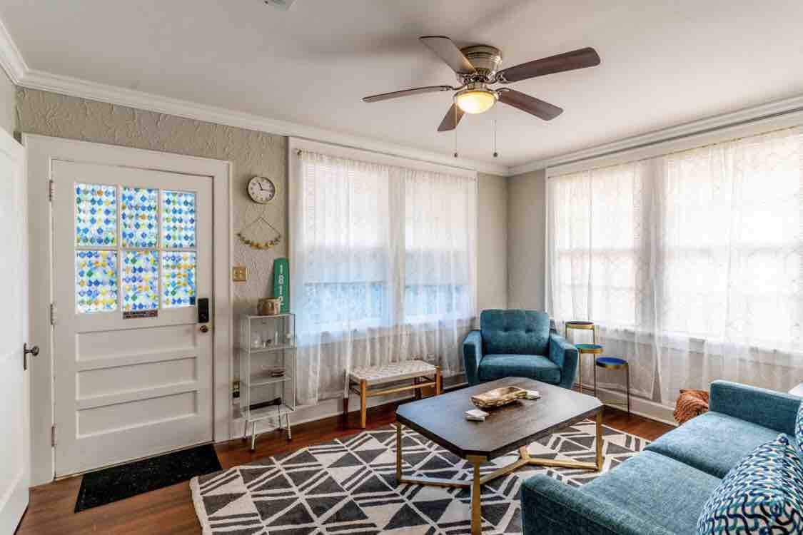 A bright living area is enhanced by natural light streaming through large, sheer-curtained windows. A patterned rug covers the hardwood floor, while a comfortable blue seating arrangement is positioned around a wooden coffee table. Decorative elements include a wall clock and a colorful glass entry door.