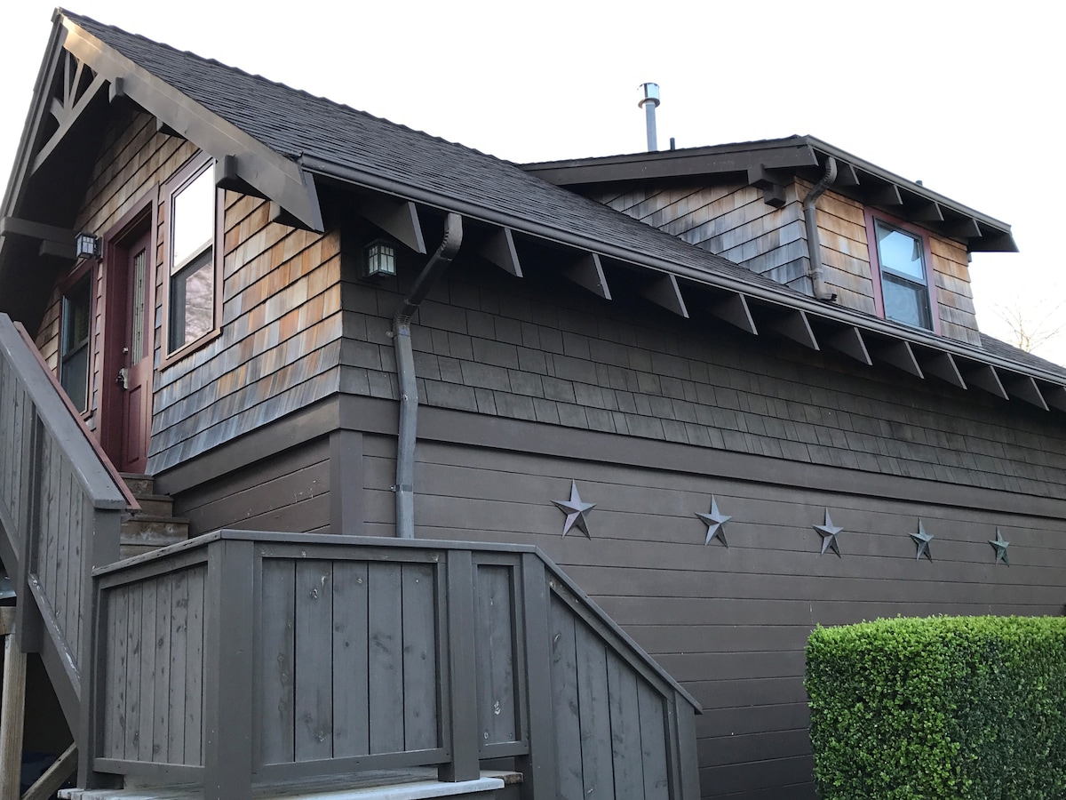 The exterior of the cottage showcases a craftsman style with wooden shingles and decorative star accents along the lower wall. A set of stairs leads up to the entrance, complemented by a porch area framed by neatly trimmed greenery.