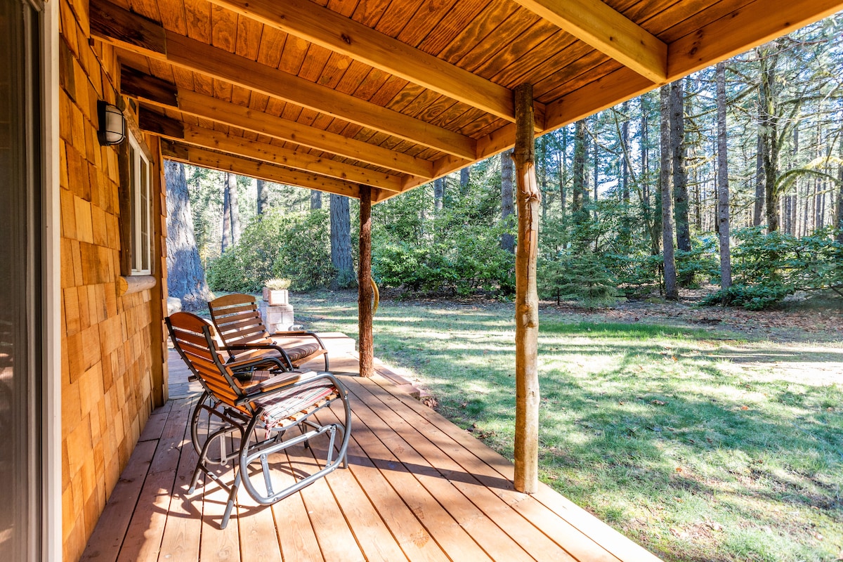 A private covered back deck features two rocking chairs positioned on a wooden floor. Tall trees and lush greenery create a serene backdrop, inviting relaxation in a peaceful outdoor setting.