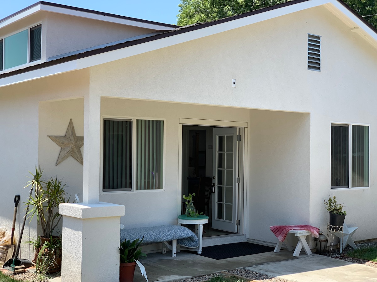 The exterior of the cottage displays a contemporary design with a light stucco finish. A welcoming entrance features a set of double doors framed by large windows, providing ample natural light. Potted plants and decorative items enhance the inviting appearance of the front porch.