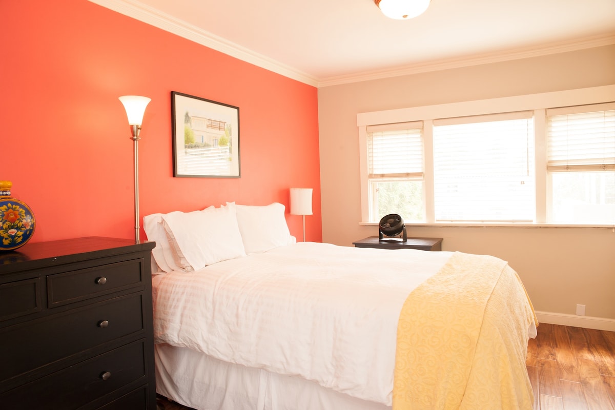 A sunny bedroom is highlighted by a vibrant red accent wall. A spacious bed with white linen is positioned beside a large dresser. Natural light filters through the windows, which are partially covered by blinds. A lamp stands next to the bed, creating a functional space.