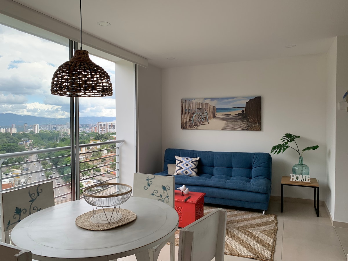 A contemporary living space features a blue sofa facing a large window, offering expansive views of the city. A round table with decorative chairs and a red accent table are present, while a woven pendant light hangs overhead, adding texture to the bright, airy environment.