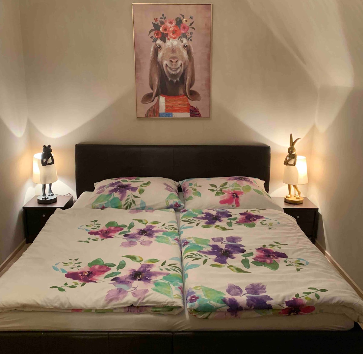 A spacious bed with floral-patterned bedding is centered against a light-colored wall. Soft bedside lamps illuminate the area, complementing a framed artwork featuring a goat adorned with flowers. The overall ambiance is calm and inviting.